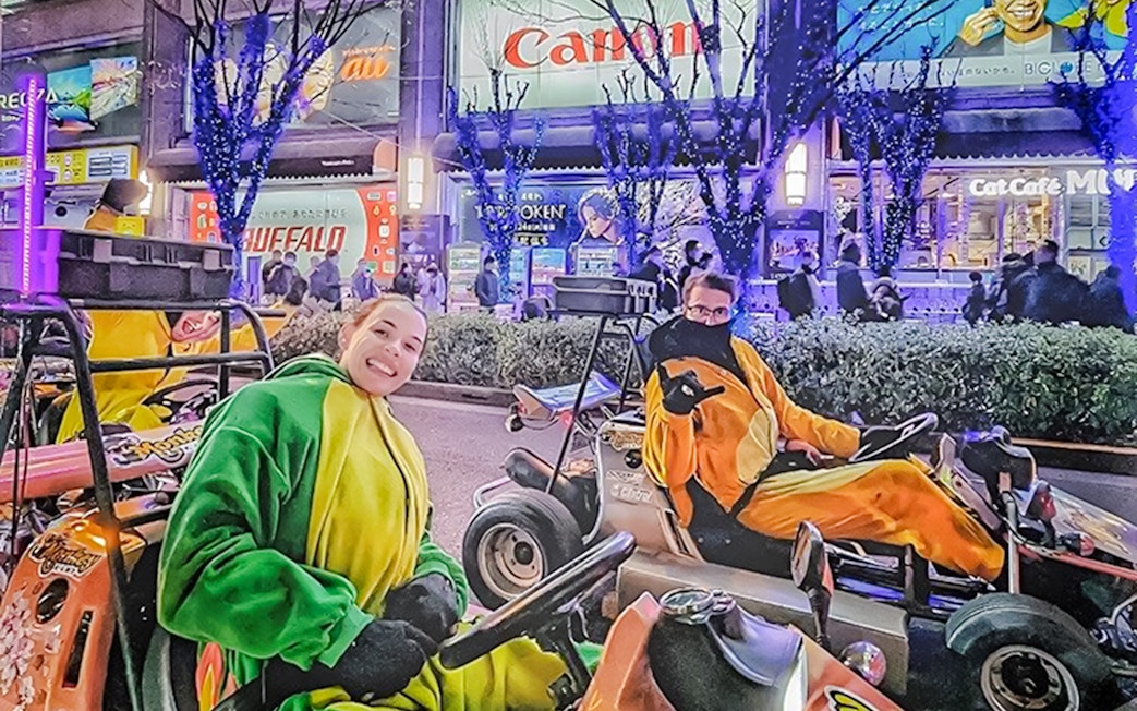 People in costume drive go-karts on a brightly lit street in Tokyo at night.