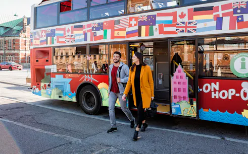 Hop-On Hop-Off bus with international flags, passengers exiting in a city setting.