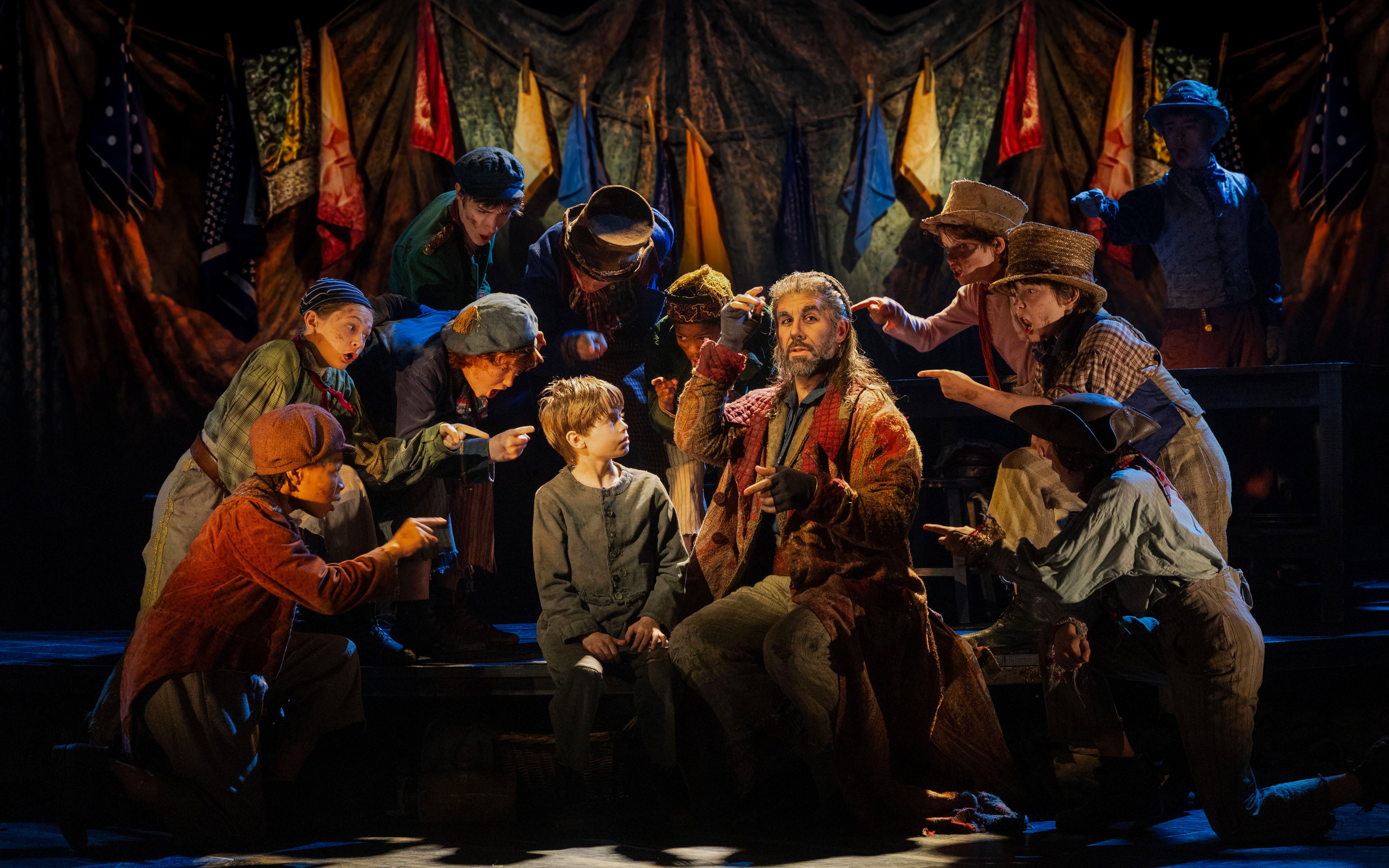Actors in period costumes perform a scene from "Oliver" on stage with colorful flags in the background.