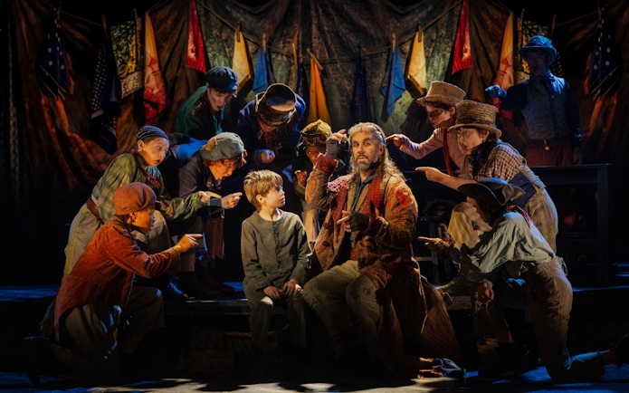 Actors in period costumes perform a scene from "Oliver" on stage with colorful flags in the background.