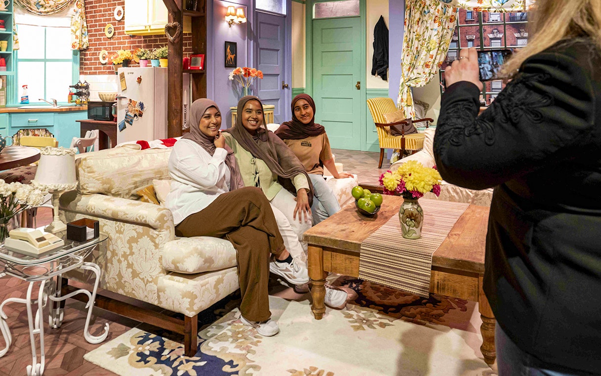 Visitors posing on the iconic FRIENDS™ set couch with kitchen backdrop.