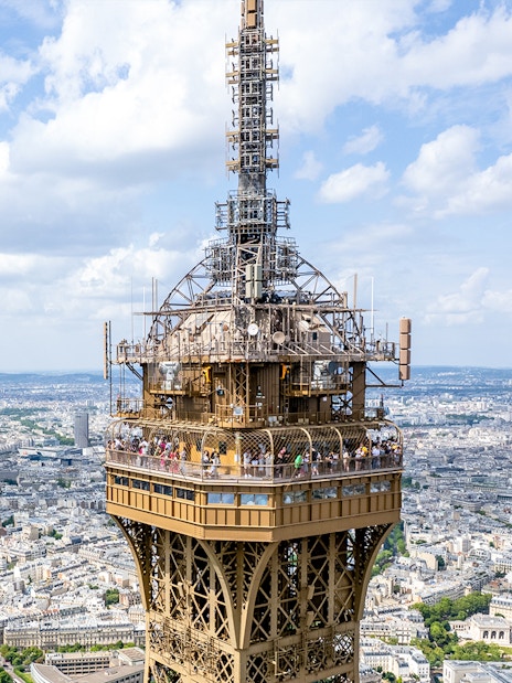Eiffel Tower summit view with tourists on the observation deck, Paris skyline in background.