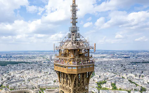 Eiffel Tower summit view with tourists on the observation deck, Paris skyline in background.