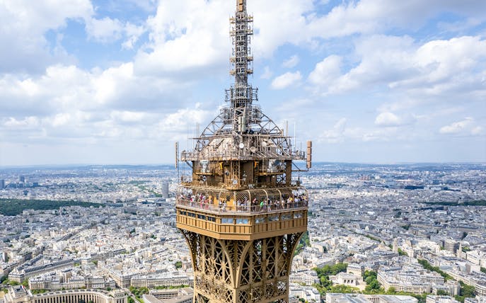 Eiffel Tower summit view with tourists on the observation deck, Paris skyline in background.