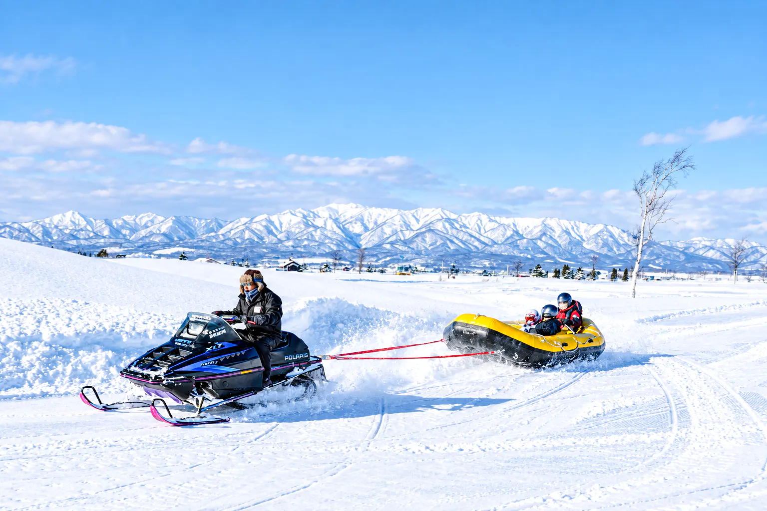 Snowmobile towing a yellow inflatable raft with riders in snowy Hokkaido landscape, Sapporo tour.