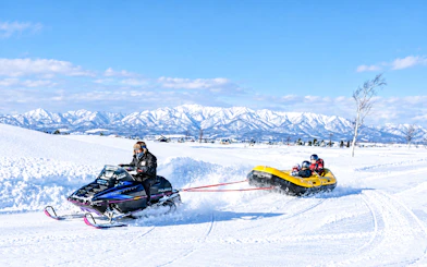 Snowmobile towing a yellow inflatable raft with riders in snowy Hokkaido landscape, Sapporo tour.