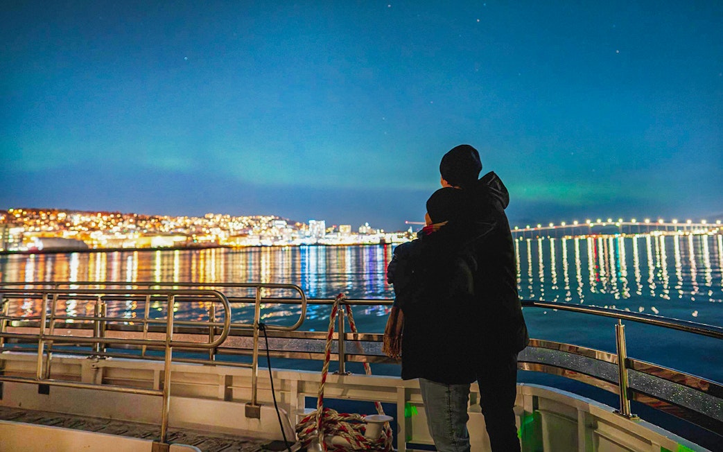 Couple on a boat viewing Northern Lights over Tromsø cityscape.