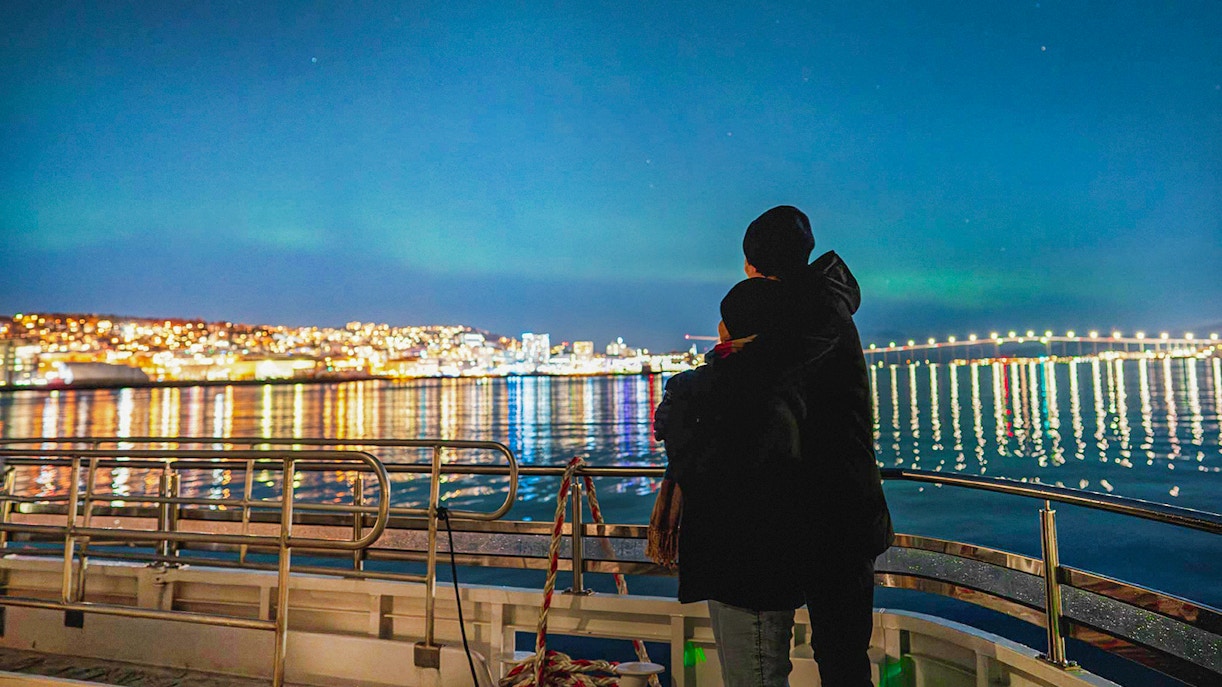Couple on a boat viewing Northern Lights over Tromsø cityscape.