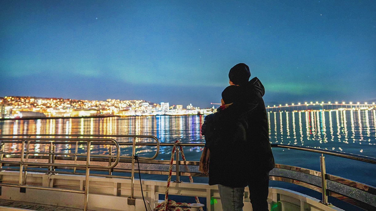 Couple on a boat viewing Northern Lights over Tromsø cityscape.