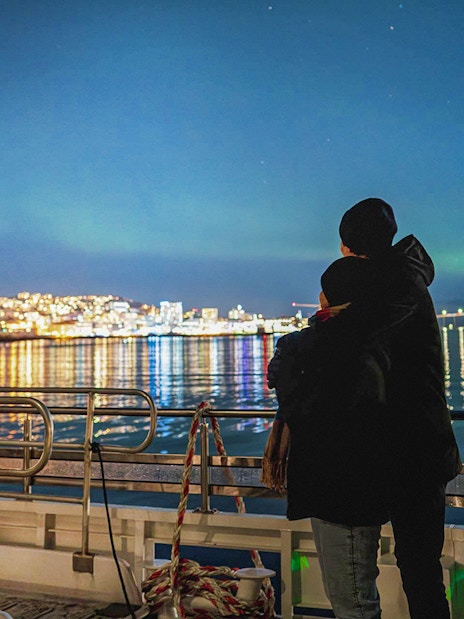 Couple on a boat viewing Northern Lights over Tromsø cityscape.