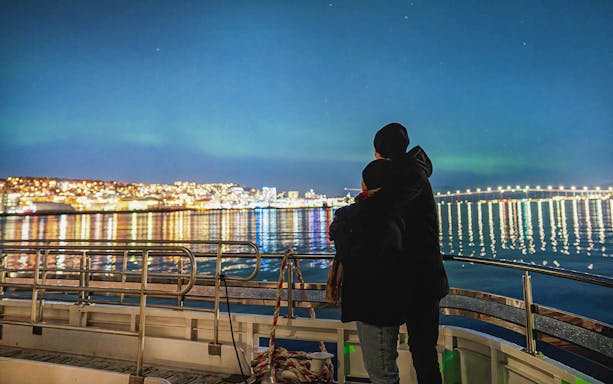 Couple on a boat viewing Northern Lights over Tromsø cityscape.