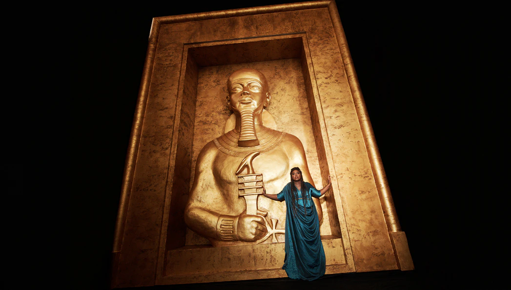 Opera performer in blue dress stands before large golden Egyptian statue in Aida production.