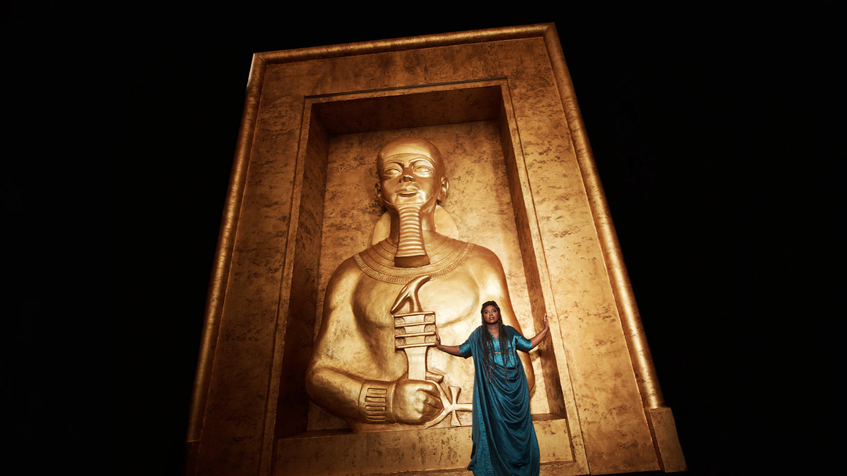 Opera performer in blue dress stands before large golden Egyptian statue in Aida production.