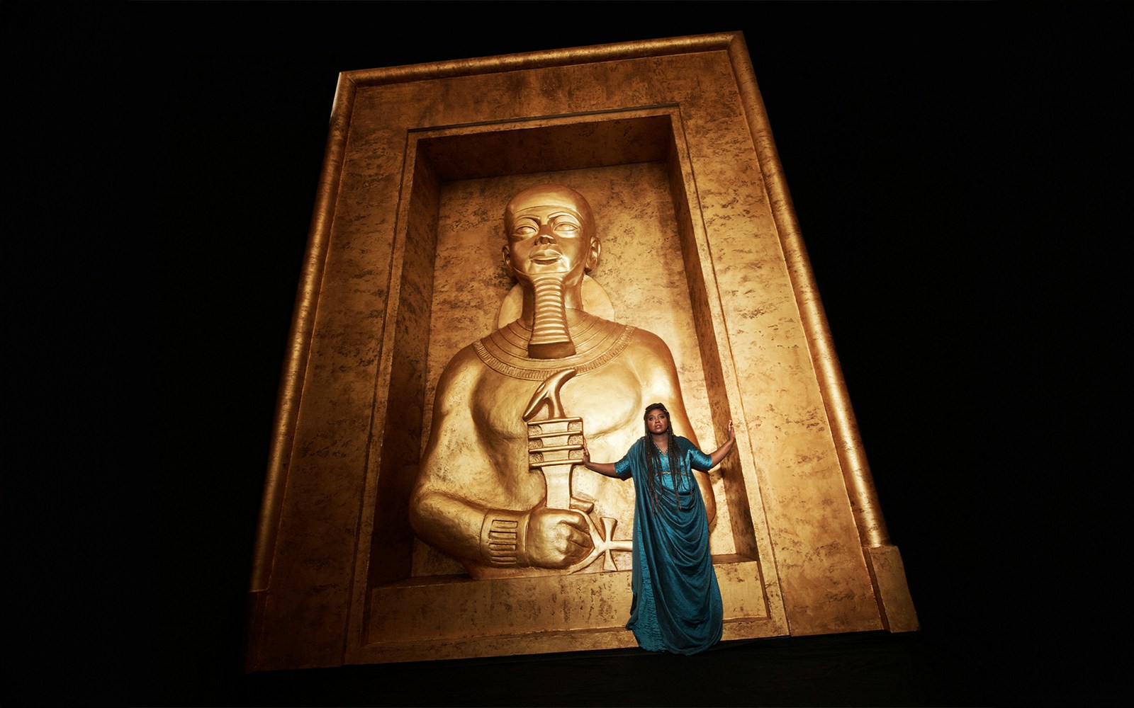 Opera performer in blue dress stands before large golden Egyptian statue in Aida production.