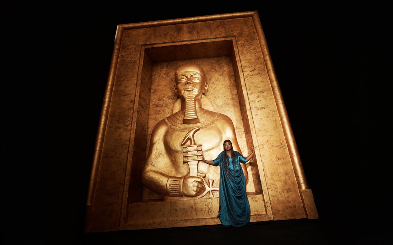 Opera performer in blue dress stands before large golden Egyptian statue in Aida production.