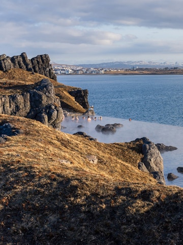 People relaxing in geothermal waters at Sky Lagoon, Iceland, with rocky coastline and distant city view.
