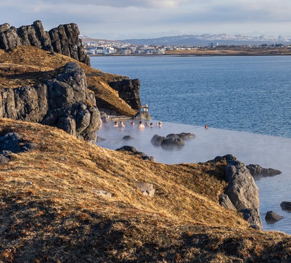 People relaxing in geothermal waters at Sky Lagoon, Iceland, with rocky coastline and distant city view.