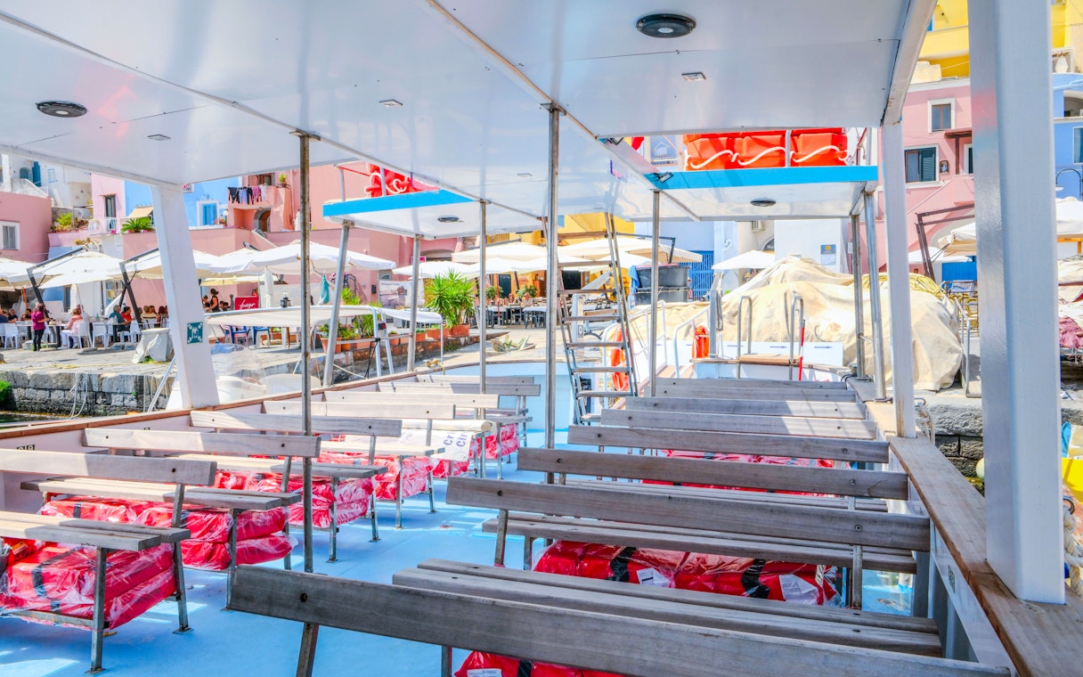 Boat deck with seating, docked at Procida Island, colorful buildings and cafes in the background.
