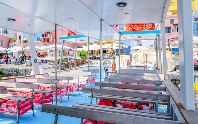Boat deck with seating, docked at Procida Island, colorful buildings and cafes in the background.