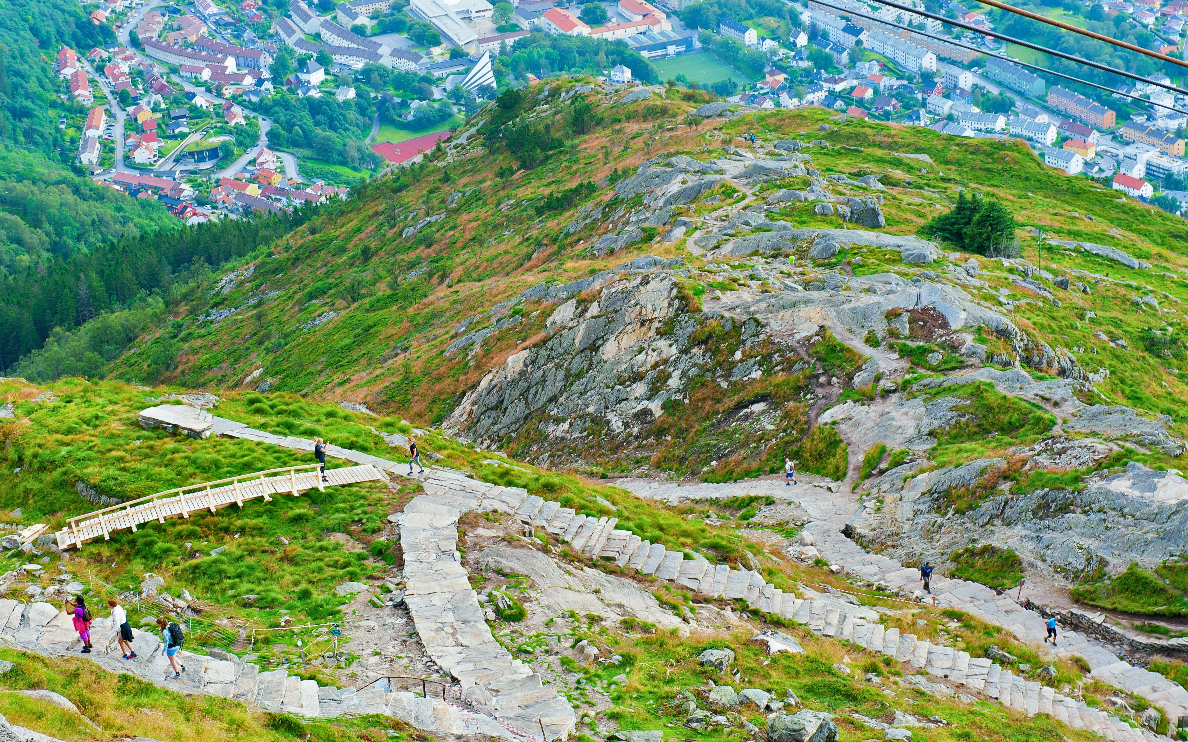 Hikers ascending stone path on Mount Ulriken with Bergen cityscape below, Norway.