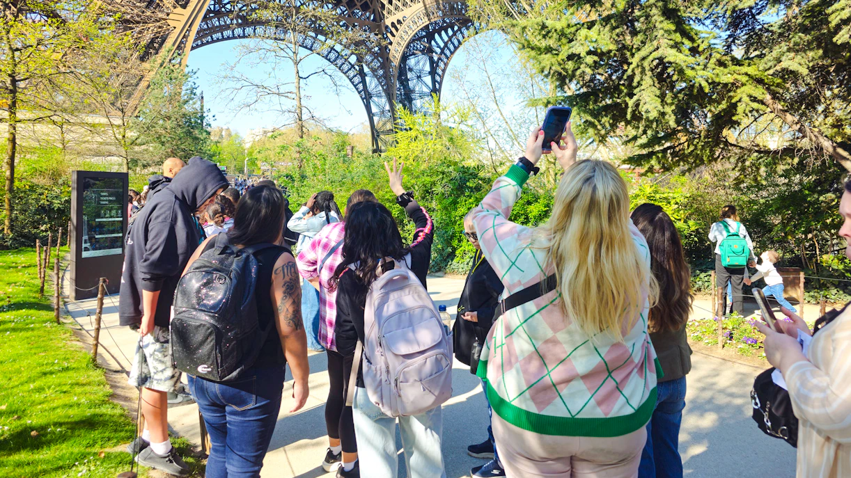 Tourists gather under the Eiffel Tower in Paris, France, capturing photos and enjoying the view.
