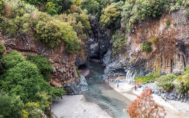 River flowing through rocky gorge at Gole Alcantara with people trekking along the banks.