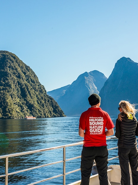 Passenger with nature guide on Milford Sound cruise, viewing scenic fjords and mountains.
