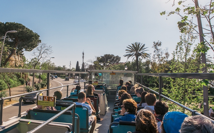 Barcelona hop-on hop-off bus with tourists, palm trees, and city street.