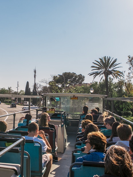 Barcelona hop-on hop-off bus with tourists, palm trees, and city street.