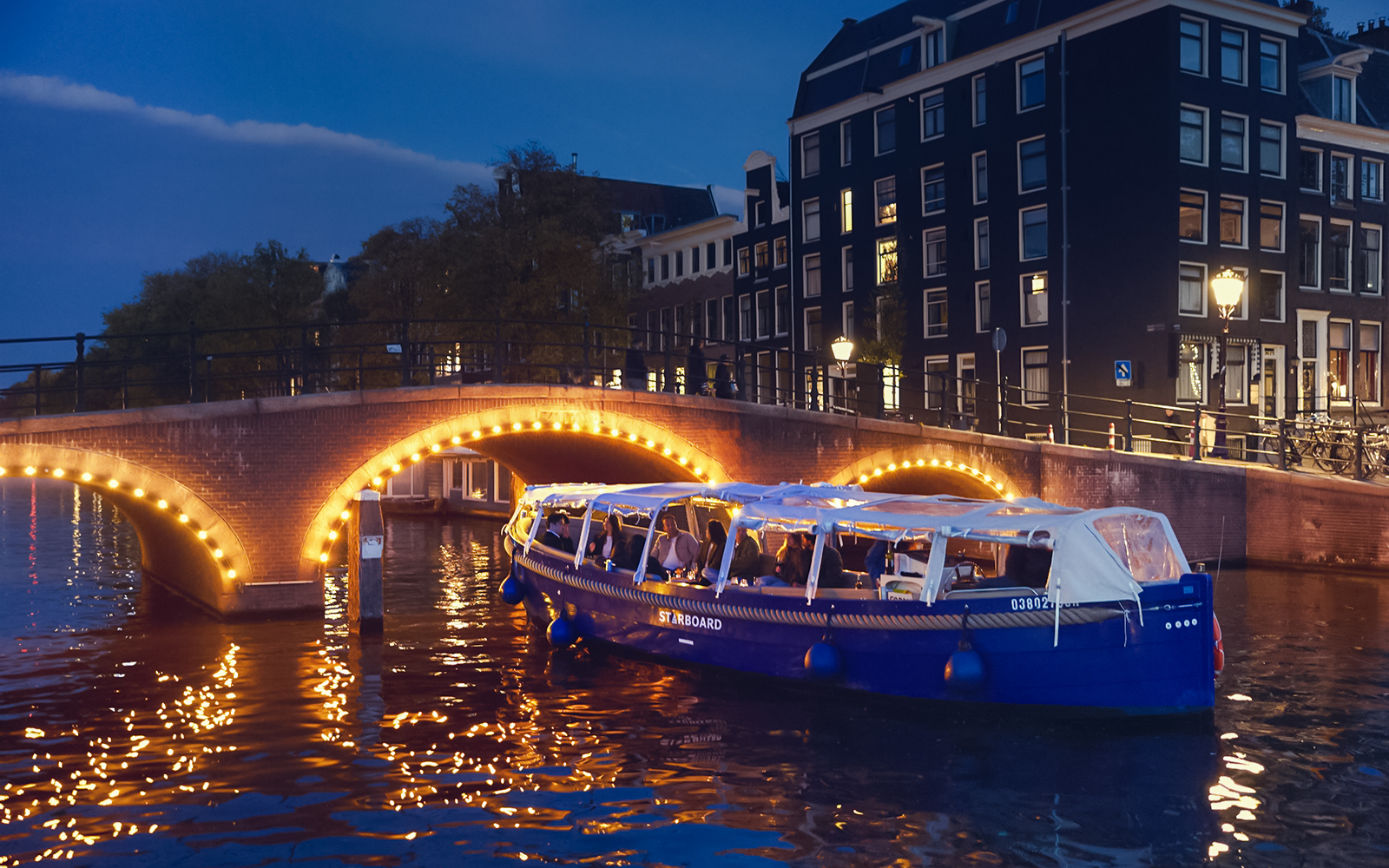 Amsterdam evening canal cruise with passengers enjoying wine and cheese under illuminated bridge.