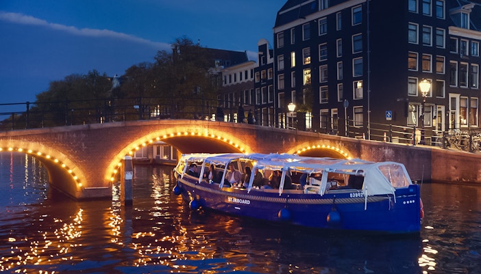 Amsterdam evening canal cruise with passengers enjoying wine and cheese under illuminated bridge.