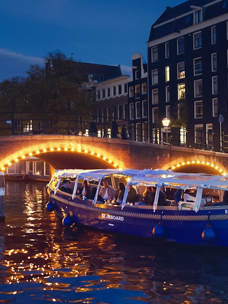 Amsterdam evening canal cruise with passengers enjoying wine and cheese under illuminated bridge.