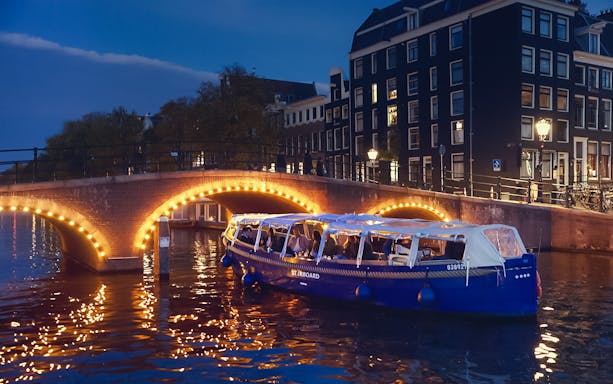 Amsterdam evening canal cruise with passengers enjoying wine and cheese under illuminated bridge.