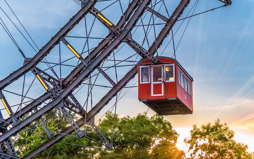 Red cabin on a giant Ferris wheel at sunset, surrounded by trees.