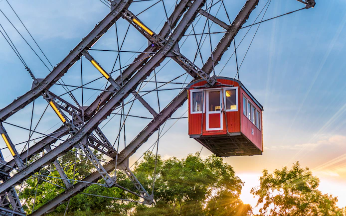 Red cabin on a giant Ferris wheel at sunset, surrounded by trees.