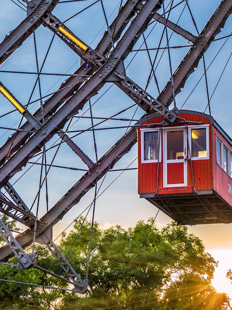 Red cabin on a giant Ferris wheel at sunset, surrounded by trees.