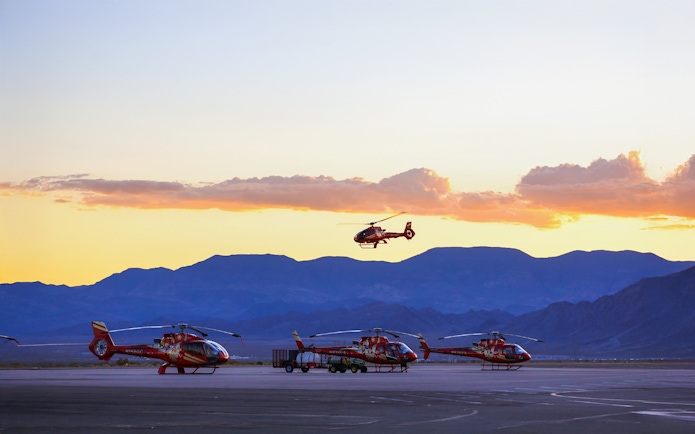 Helicopters parked on tarmac with one in flight at sunset, mountains in background.
