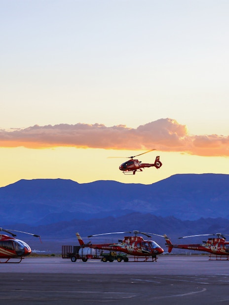 Helicopters parked on tarmac with one in flight at sunset, mountains in background.