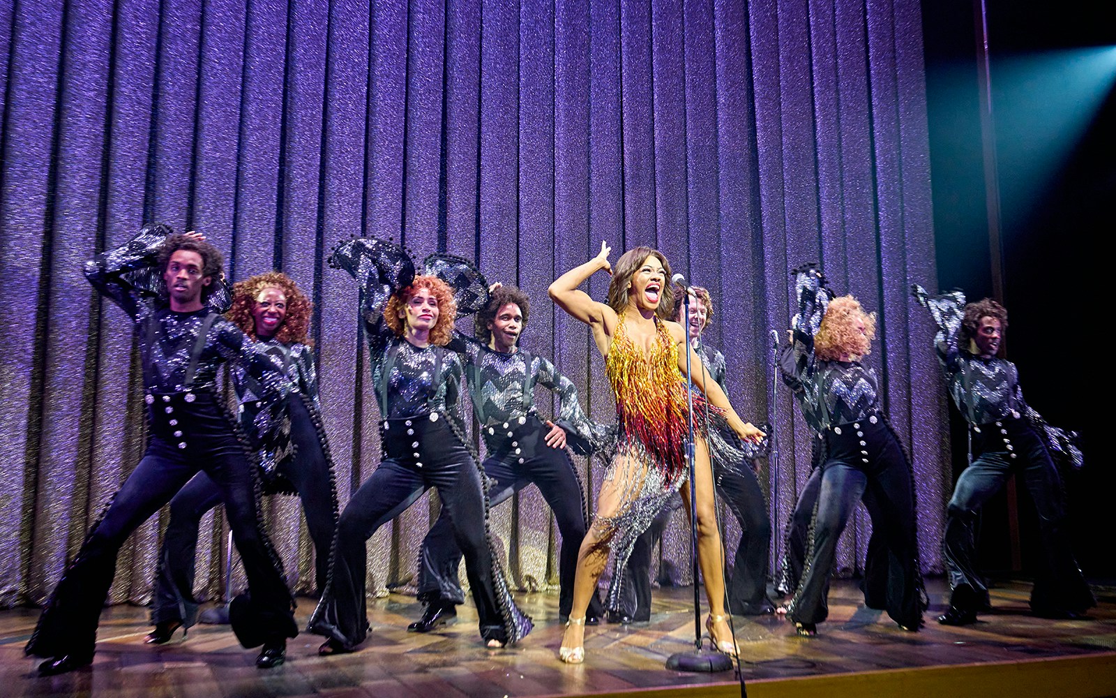 Performers on stage during TINA: The Tina Turner Musical, with lead singer and dancers in vibrant costumes.