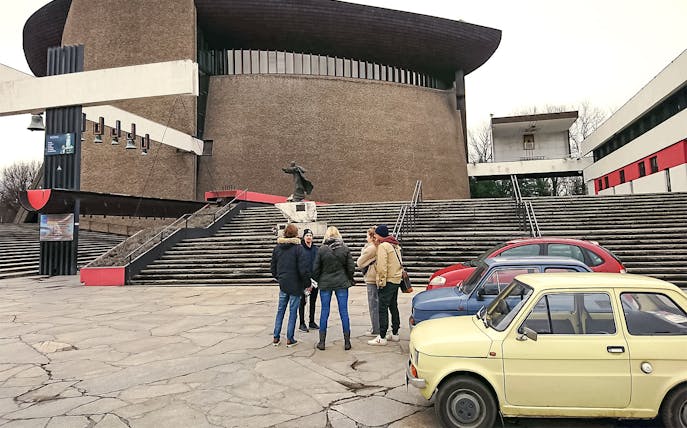 Group touring Nowa Huta in Kraków with retro cars parked nearby.