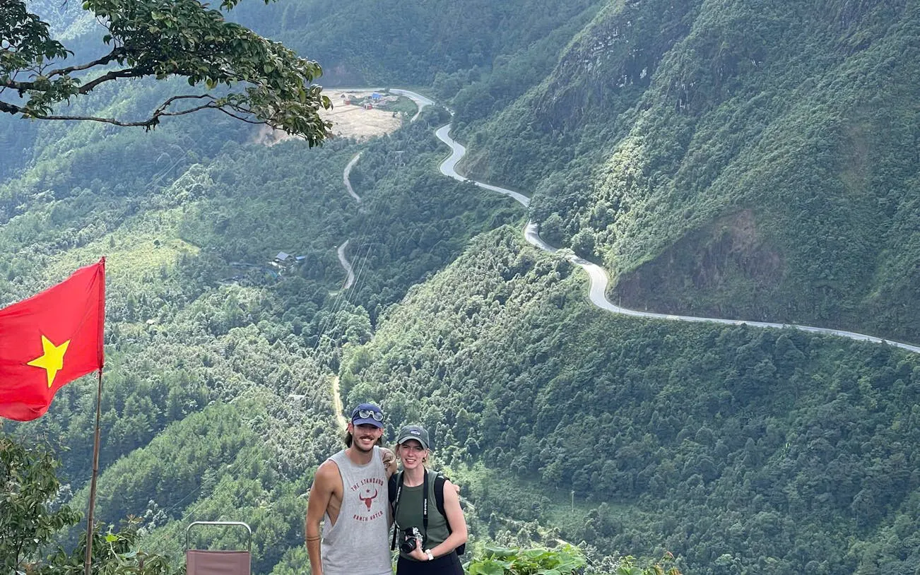 Couple posing with Vietnamese flag overlooking winding mountain road in Sapa on motorbike tour.