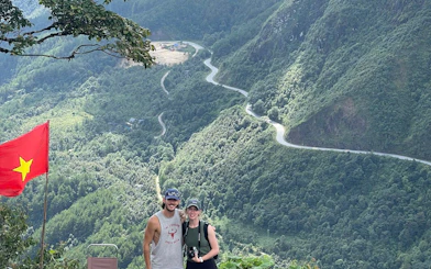 Couple posing with Vietnamese flag overlooking winding mountain road in Sapa on motorbike tour.
