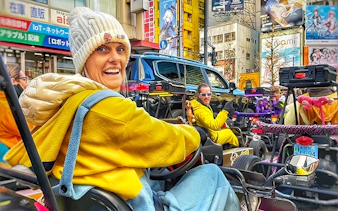 Woman in a go-kart on a busy Tokyo street, wearing a white beanie and yellow hoodie.