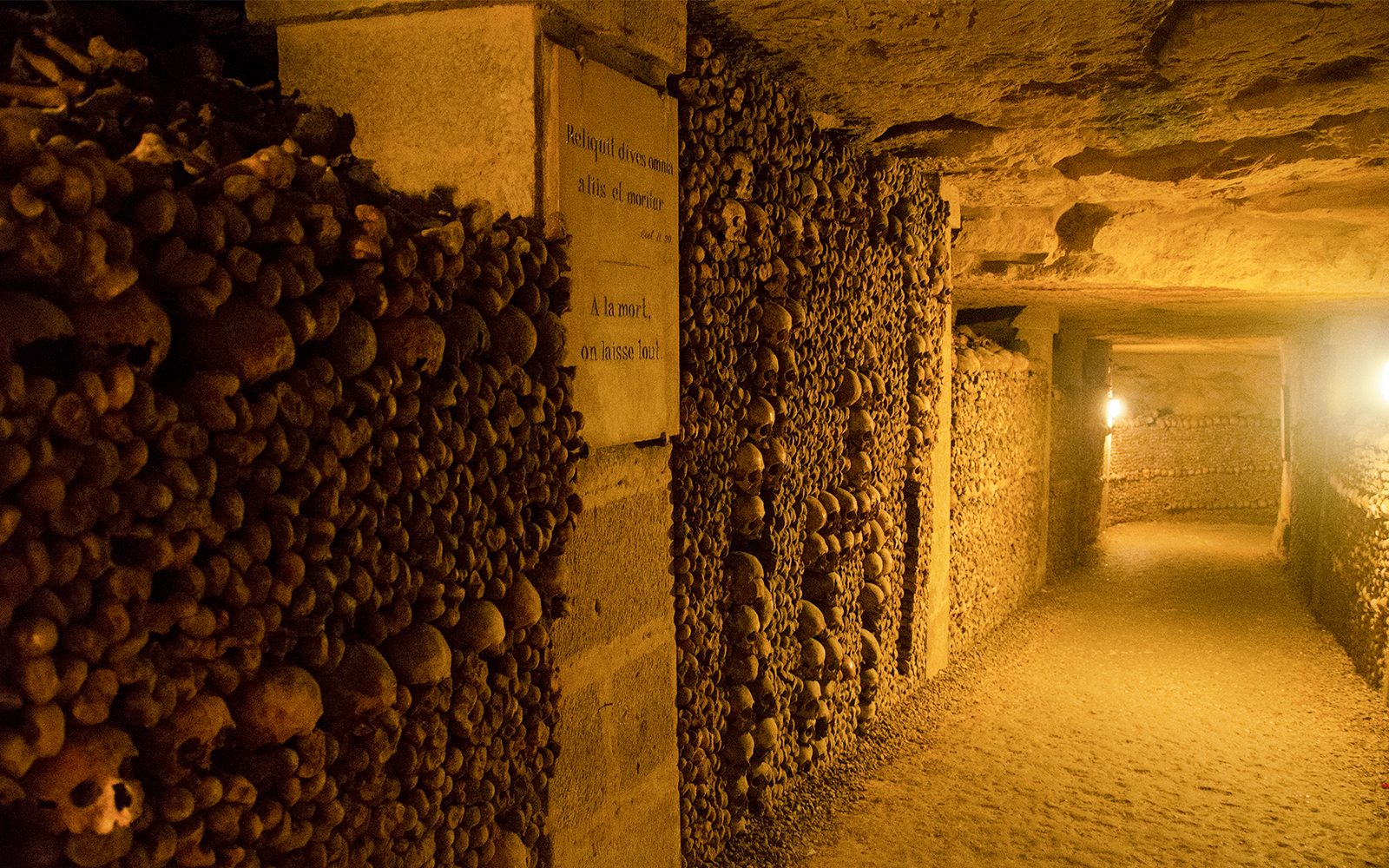 Paris Catacombs corridor lined with skulls and bones, dimly lit passageway.