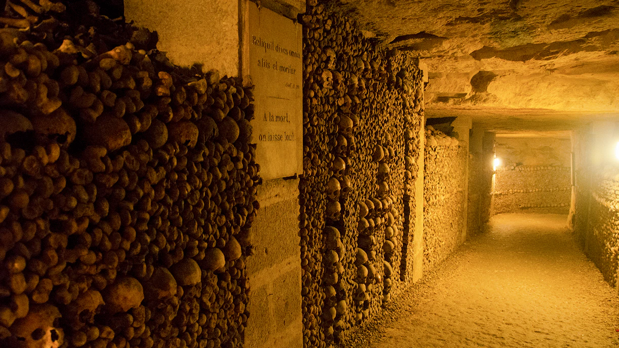 Paris Catacombs corridor lined with skulls and bones, dimly lit passageway.