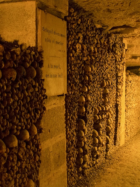 Paris Catacombs corridor lined with skulls and bones, dimly lit passageway.