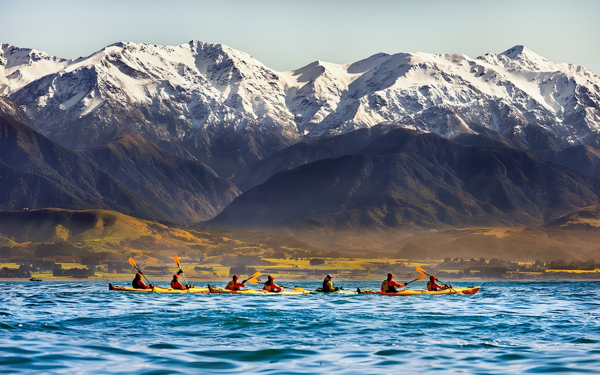 Kayakers paddling on a lake with snow-capped mountains in the background.