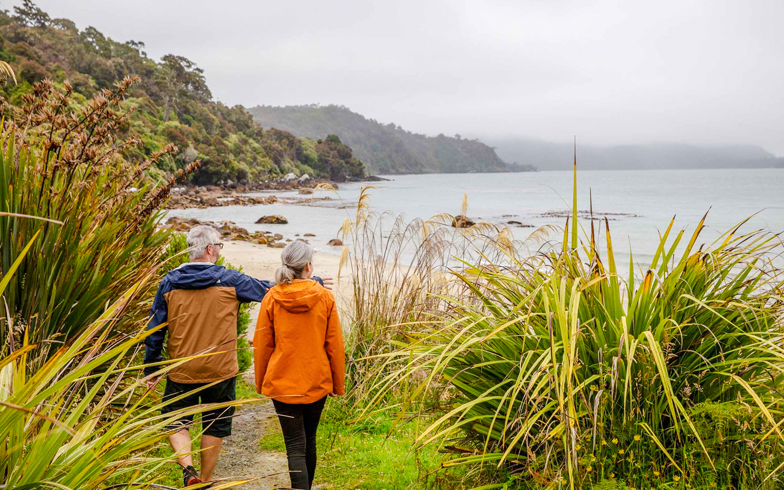 Couple walking along a coastal path on Stewart Island, surrounded by lush greenery and ocean views.