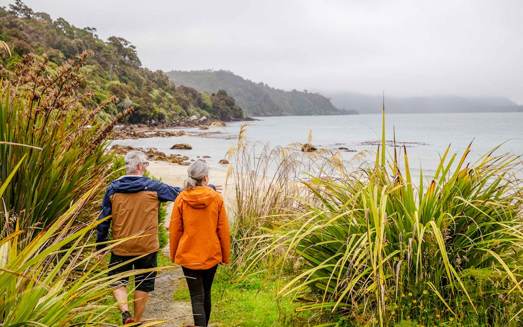 Couple walking along a coastal path on Stewart Island, surrounded by lush greenery and ocean views.