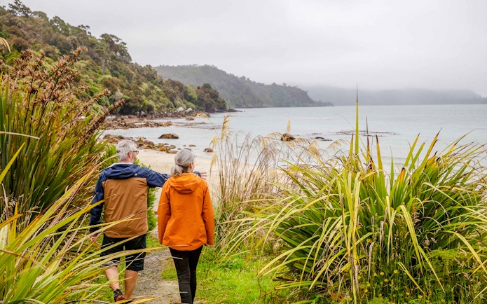 Couple walking along a coastal path on Stewart Island, surrounded by lush greenery and ocean views.